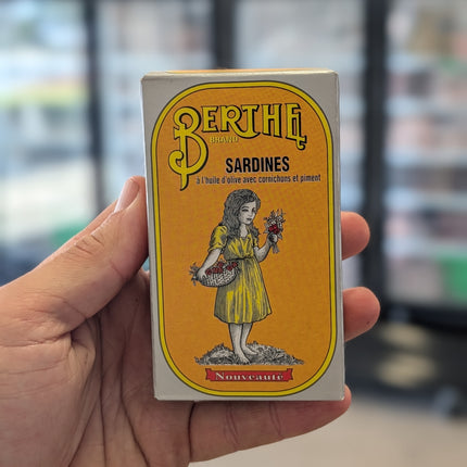 Hand holding a vintage-style Berthe sardine can packaging with a yellow label.