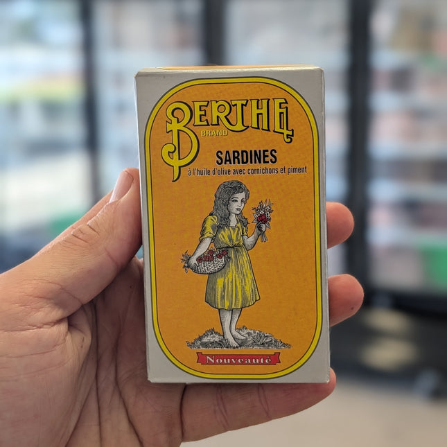 Hand holding a vintage-style Berthe sardine can packaging with a yellow label.