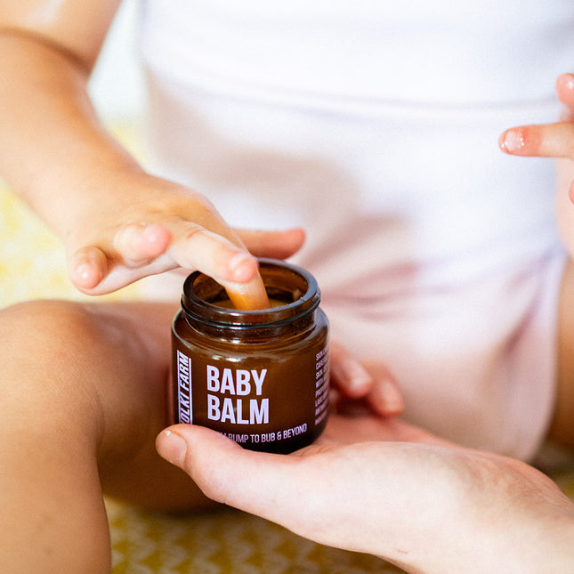 Person holding a jar of baby balm with a child's hand reaching towards it.