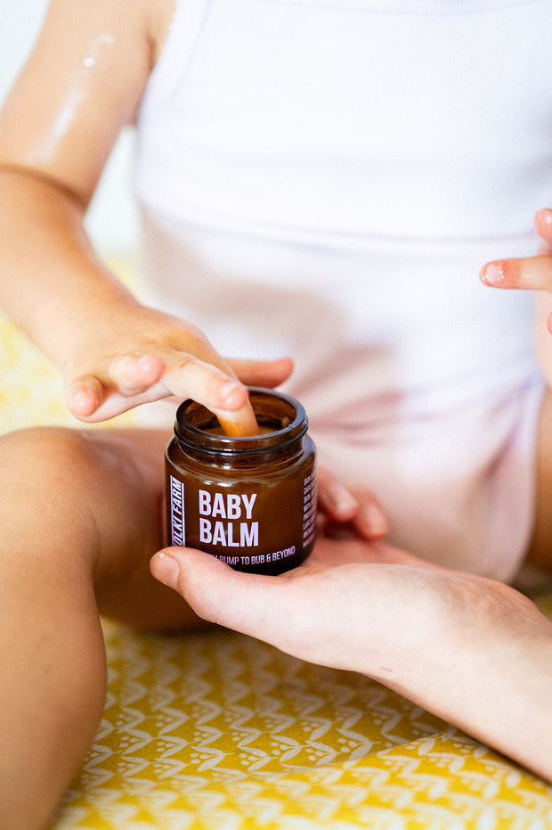 Person holding a jar of baby balm with a child's hand reaching towards it.
