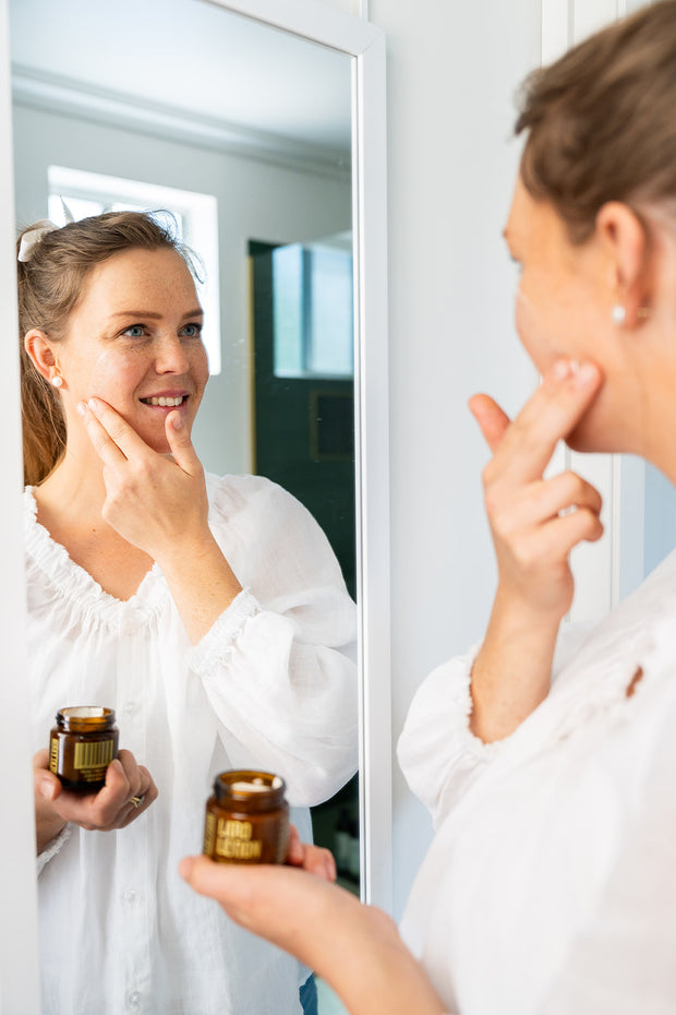 Woman applying a skincare product to her face in front of a mirror, holding jars of the product.