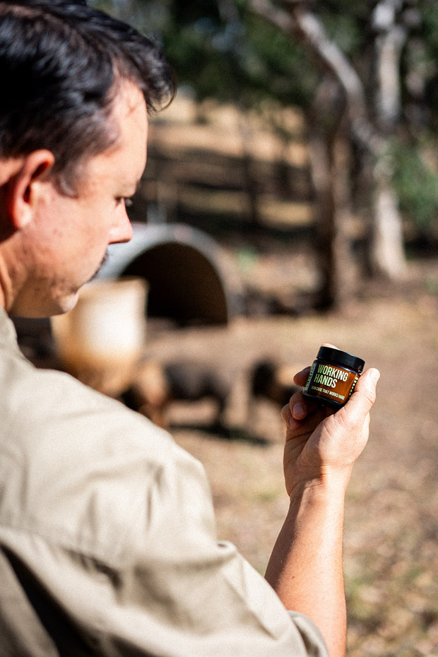 Man holding a jar of working hands lard balm outdoors with trees and a barrel in the background